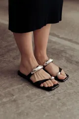 Sandalias negras de cuero con tira ancha sobre el empeine y tira entre los dedos, ambas adornadas con apliques metálicos plateados.