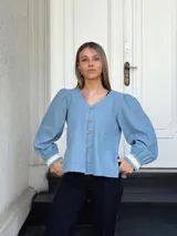Camisa celeste de jean con cuello en V, mangas largas abullonadas con puños de encaje y cierre frontal con botones.