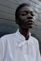 Camisa blanca de popelín con cuello redondo con volante y lazo, manga larga y bajo peplum. Cierre frontal con botones.