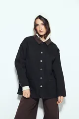 Sobrecamisa tipo barn jacket color negro, de algodón con elastano, con silueta relajada y estructura firme. Presenta cierre frontal con botones metálicos, cuello marrón, bolsillos aplicados y pespuntes visibles en color marrón.