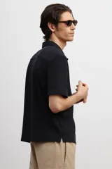 Camisa negra de punto con textura, de corte recto, manga corta, cuello inglés, tapeta lisa, canesú en la espalda y bajo recto con aberturas laterales.