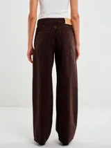 Pantalón de denim color marrón oscuro, tiro medio, con pierna ancha y recta. Presenta diseño clásico de cinco bolsillos y cierre con botón y cremallera.