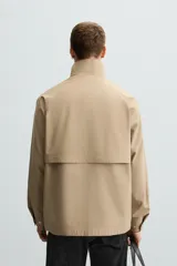 Parka ligera color beige, de corte relaxed fit, confeccionada en tejido técnico repelente al agua. Tiene cuello alto, mangas largas con puños ajustables con botón a presión, bolsillos de solapa en la cadera y cierre frontal con cremallera oculta por solapa con botones a presión.