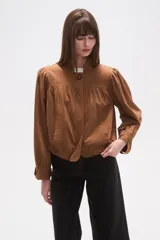 Campera marrón de manga larga con cuello mao y cierre frontal de un solo botón. Presenta pinzas y frunces en el canesú delantero y en los hombros, creando un efecto abullonado en las mangas. El bajo es ajustado con elástico.