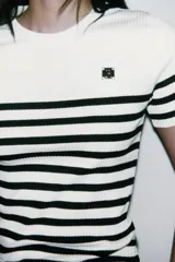 Camiseta de rib a rayas horizontales blancas y negras, de corte ajustado, cuello redondo y manga corta. Presenta un broche metálico en el frente y una medalla con el logo de Zara en la espalda.