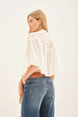 Camisa blanca de algodón con cuello redondo, corte amplio y mangas cortas abullonadas. Presenta detalles de calado en la parte superior delantera y trasera. Cierre frontal con botones.