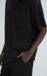 Camisa negra de manga corta estilo técnica, confeccionada en microfibra liviana. Tiene doble bolsillo frontal con cierre de cremallera y broches en lugar de botones.