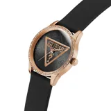 Reloj Guess con caja de metal color oro rosado, bisel decorado con cristales y correa de silicona negra. La esfera es negra con patrón de monograma y un triángulo central con el logo de Guess.