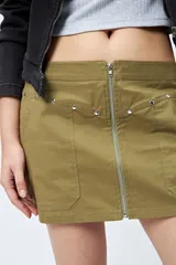 Minifalda color marrón claro, de corte recto con cierre metálico visible al frente y bolsillos decorativos con tachas.