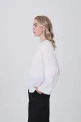 Blusa blanca de poliéster, entallada, con escote en V y botones frontales. Presenta mangas largas con puños anchos.