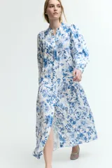 Vestido largo blanco con estampado floral en tonos azules. Tiene cuello mao, botones ocultos en la parte delantera, mangas largas abullonadas con puños elásticos, cinturón fino extraíble y costura fruncida debajo de la cadera para dar vuelo.