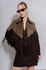 Chaqueta tipo bomber de cuero ovino desgastado marrón claro con detalles en gamuza marrón oscuro. Presenta un canesú en pico con flecos largos en el frente y espalda, cuello solapa y cierre frontal con broches forrados.