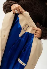 Trench largo de cuero color beige con forro azul, cuello clásico, cierre frontal con botones y bordado de las "S" en el frente.