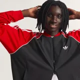 Campera Adidas Originals de corte holgado, con cuerpo en tejido de malla negro y mangas en color rojo con las icónicas tres tiras blancas. Presenta cierre frontal completo, cuello tipo bomber y logo del trébol bordado en el pecho.