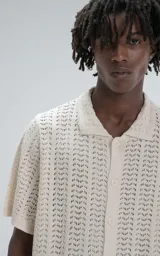 Camisa de tejido de punto color beige, manga corta, con cuello polo y cierre frontal de botones. Presenta un calce regular y un patrón calado en zigzag.