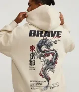 Hoodie color beige claro, corte boxy, con capucha ajustable y bolsillo canguro. Presenta un gran estampado gráfico en la espalda con un dragón oriental, texto en inglés y caracteres japoneses, y la palabra "BRAVE" en la parte superior.