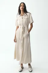 Vestido camisero largo color beige, de manga corta y cuello solapa. Tiene bolsillos de parche en el pecho, bolsillos laterales ocultos y cinturón de la misma tela.