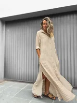 Vestido camisero largo con estampado de rayas verticales en beige y blanco. Presenta cuello camisero, cierre frontal con botones, mangas largas y tajos laterales en el ruedo.