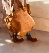 Bolso tote color marrón claro con textura símil piel de cocodrilo. Tiene doble asa corta, laterales de gamuza con flecos y un aplique floral en el frente.