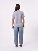 Remera blanca de algodón con cuello redondo y manga corta. Presenta un pequeño bordado en el pecho izquierdo con una figura que parece ser una persona acostada en una tabla de surf o flotador.