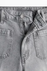 Pantalón de jean de tiro alto, color gris lavado, con pierna ancha y puños doblados. Presenta bolsillos de parche grandes en la parte delantera y bolsillos traseros.