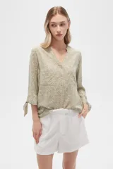 Blusa color beige con estampado de arabescos blancos, cuello mao, mangas 3/4 con lazos en los puños, cierre parcial con botones y bolsillos frontales.