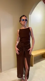 Vestido largo marrón oscuro, ajustado al cuerpo, con escote cuadrado y tirantes finos. La falda presenta un drapeado frontal anudado que crea una abertura alta.