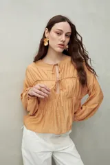 Blusa color beige de caída fluida con cuello redondo y lazos frontales. Diseño con cortes horizontales y frunces, mangas largas con puños elastizados.