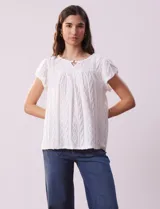 Blusa blanca labrada con cuello a la base, escote en V y aplique metálico. Tiene mangas cortas superpuestas.
