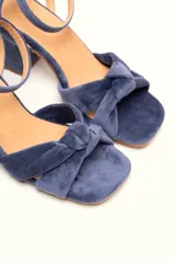 Sandalia de terciopelo verde con taco cuadrado de 7 cm, punta cuadrada y tiras anchas anudadas en el empeine. Cuenta con una tira fina ajustable al tobillo con hebilla dorada.