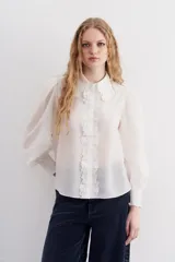 Camisa blanca de algodón con corte recto, cuello clásico y cierre frontal con botones. Presenta detalles de puntilla en cuello y cartera, mangas largas abullonadas con puños elastizados.