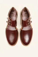Zapato tipo monk strap con aberturas laterales, confeccionado en cuero vacuno color bordó. Presenta doble tira con hebillas metálicas plateadas en el empeine y suela de cuero con taco bajo.