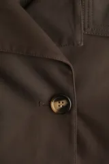 Gabardina corta color marrón chocolate, de corte holgado, con cuello clásico, doble botonadura, charreteras, trabillas abotonadas en los puños, bolsillos delanteros con solapa y pliegue invertido en la espalda. Forrada.