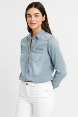 Camisa de denim celeste, corte entallado, con cuello clásico, cierre frontal con botones a presión, mangas largas con puños abotonados, dos bolsillos con solapa abotonada en el pecho y canesú en la espalda.