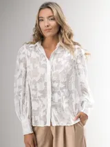 Camisa blanca con estampado floral, cuello clásico, cierre frontal con botones y mangas largas abullonadas con puños abotonados.