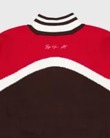 Campera de tejido de punto de grosor medio, con cuerpo marrón oscuro, mangas rojas y detalles en blanco en los hombros, cuello y puños. Presenta cierre frontal y un pequeño logo bordado en el pecho.