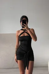 Vestido corto negro ajustado, con escote strapless y tirantes finos que se cruzan en el cuello con un anillo central. Presenta frunces laterales y un lazo anudado en la cintura.
