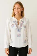 Blusa blanca con bordados florales en tonos celeste, rojo y marrón. Tiene cuello redondo con lazo ajustable y mangas largas abullonadas.