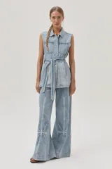 Chaleco largo de jean celeste con cuello camisero, sin mangas, con bolsillos frontales, laterales e internos, y lazo ajustable en la cintura.