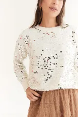 Sweater color beige de tejido de punto con cuello redondo y mangas largas. Presenta apliques de lentejuelas plateadas en el frente y las mangas.
