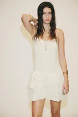 Vestido corto color crudo, de gasa, con escote en V y tirantes finos regulables. Presenta cordón cruzado con lazada en el delantero y falda con volantes. Cierre trasero con cremallera oculta.