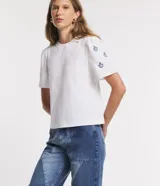 Remera blanca de algodón con cuello redondo, mangas cortas abullonadas y pequeños bordados florales azules en las mangas.