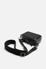 Bandolera rígida negra con formato de caja. Tiene un compartimento principal con cierre de cremallera, un bolsillo interior plano y asa de hombro ajustable y extraíble.
