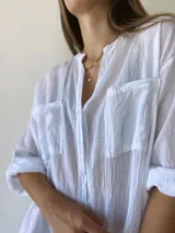 Vestido camisero blanco de corte holgado, con cuello mao, cierre frontal con botones, mangas 3/4 con puños abotonados, dos bolsillos de parche en el pecho y aberturas laterales en el ruedo.