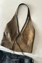 Top marrón de cuero ovino vintage con cuello halter, espalda descubierta y cierre con lazos. Presenta detalle artesanal de cutouts y pasamanería en negro alrededor del cuello.