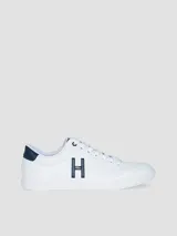 Zapatillas urbanas de corte bajo, color blanco, con cordones y suela de goma. Presentan un gran logo 'H' en color azul marino en el lateral y detalles en azul marino en el talón y la lengüeta.