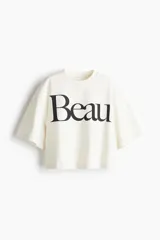 Remera cropped color crudo de corte holgado con cuello redondo acanalado, hombros caídos y bajo recto. Estampado frontal con la palabra 'Beau' en color negro.