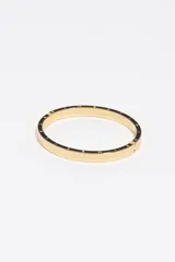 Pulsera rígida tipo brazalete de color dorado con un detalle circular de esmalte negro que presenta el logo de la marca. Cuenta con un cierre de bisagra para mayor seguridad.