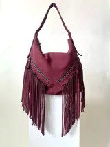 Bolso artesanal de cuero color borgoña, con flecos cortados a mano y dos bolsillos con cierre en el frente. El cerramiento principal es mediante un cierre metálico, marca YKK. Cuenta con una manija regulable con dos hebillas de metal en los extremos.