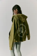 Buzo verde militar de algodón rústico con capucha, corte oversize, cierre metálico, bolsillos delanteros, estampa en frente y espalda, cordón con puntera de goma y lavado acid wash suave.